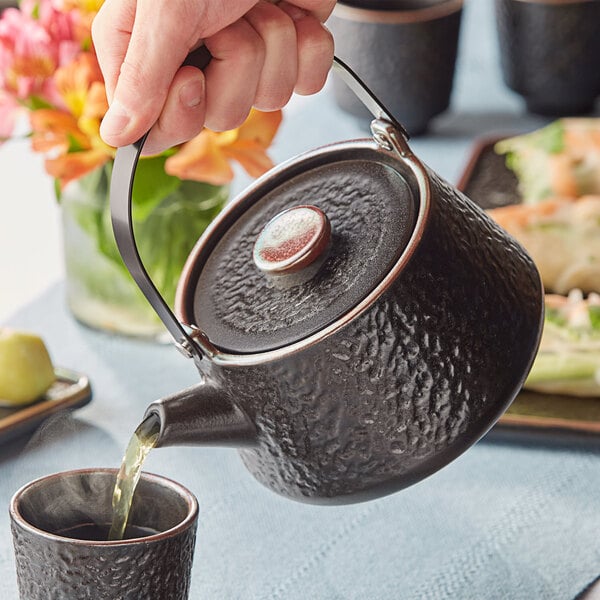 A hand pouring tea into a black Acopa stoneware teapot.