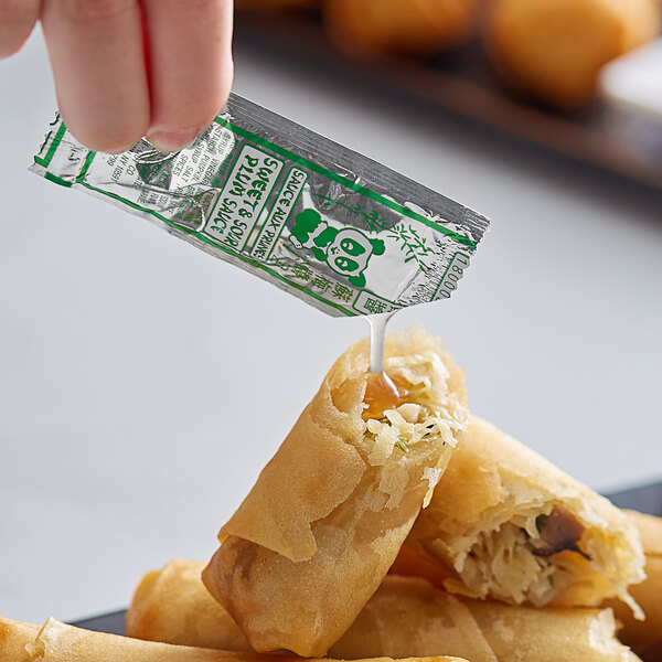A hand dipping food into a Kari-Out plum sauce packet on a table.