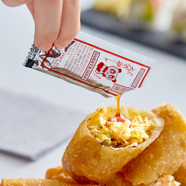 A hand dipping food into a Kari-Out sweet and sour sauce packet on a table.