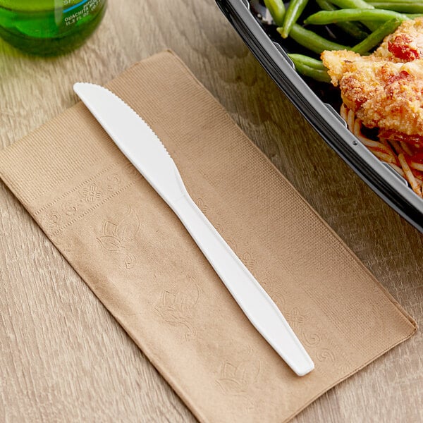 A white plastic Choice heavy weight polypropylene knife on a napkin next to a plate of food.