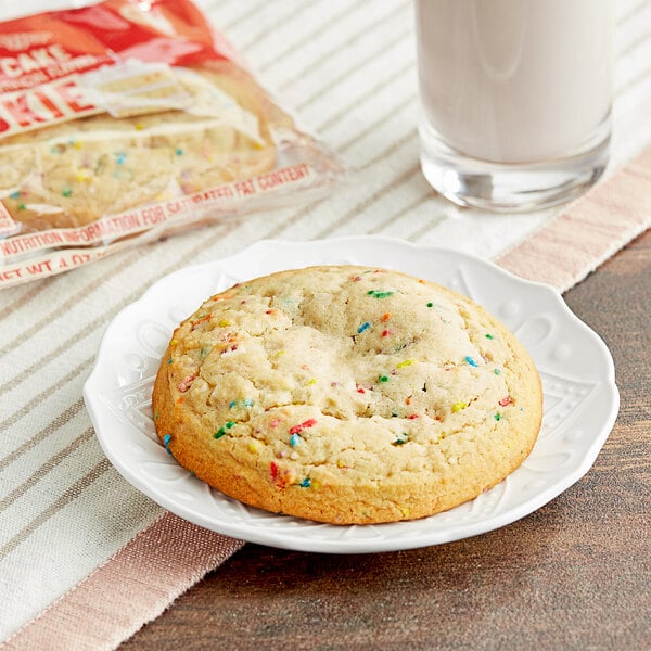 An Otis Spunkmeyer birthday cake cookie with sprinkles on a plate next to a glass of milk.