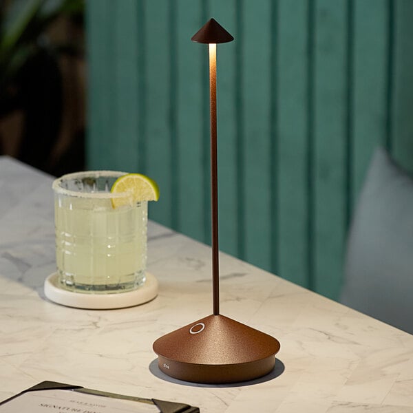 A brown Zafferano America Pina Pro cordless table lamp on a table next to a glass of liquid with a slice of lemon.