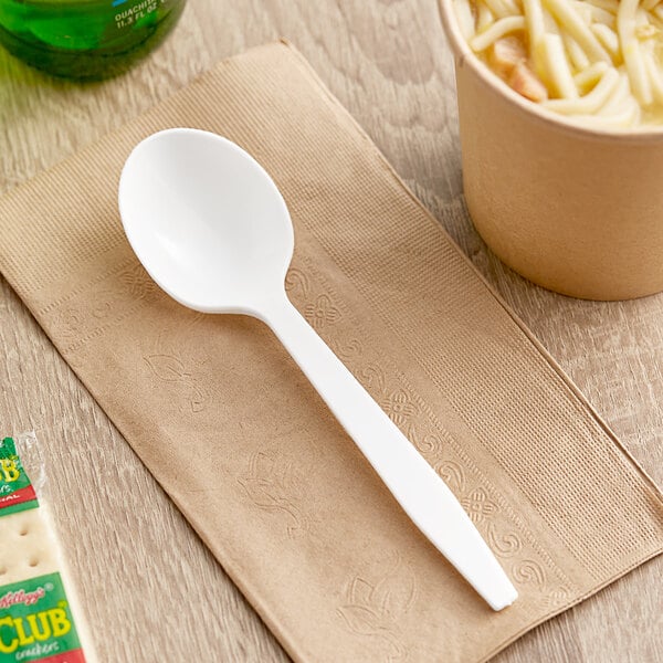 A white Choice heavy weight plastic soup spoon on a napkin next to a cup of noodles and a drink.