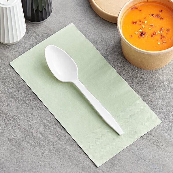 A white Choice heavy weight plastic teaspoon on a green napkin next to a bowl of soup.