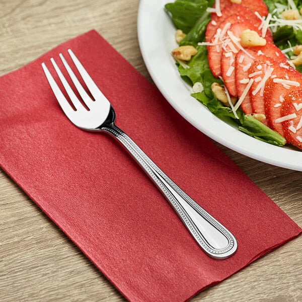 A Choice Milton stainless steel salad fork on a red napkin next to a plate of salad.