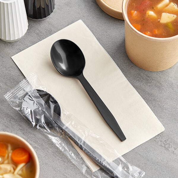 A black plastic spoon wrapped in a plastic bag next to a bowl of soup.