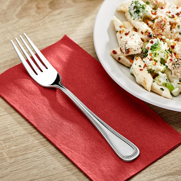 A plate of pasta on a table with a fork on a red napkin.