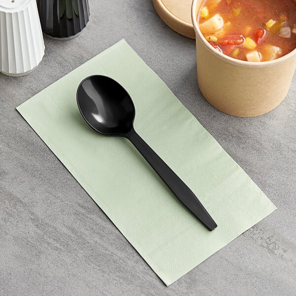 A black Choice heavy weight plastic soup spoon on a green napkin next to a bowl of soup.