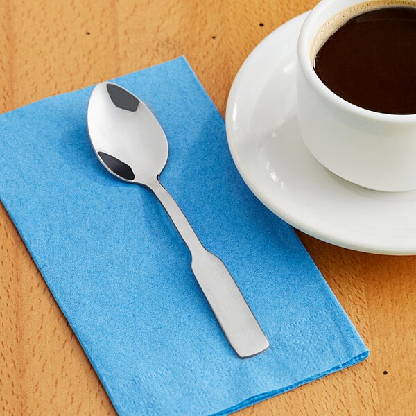 A Choice medium weight stainless steel teaspoon on a blue napkin next to a cup of coffee.