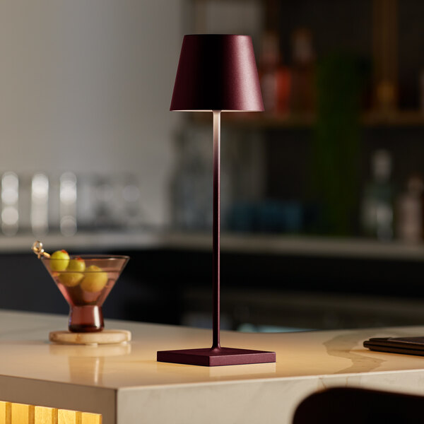 A Zafferano America Poldina Pro Bordeaux cordless table lamp on a black counter next to a martini glass with a drink and grapes.