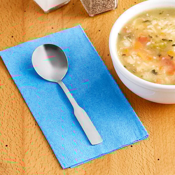 A bowl of soup and a Choice Bellwood stainless steel bouillon spoon on a napkin.