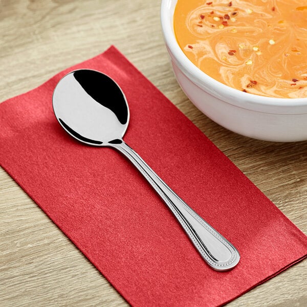 A Choice Milton stainless steel bouillon spoon on a red napkin next to a bowl of soup.