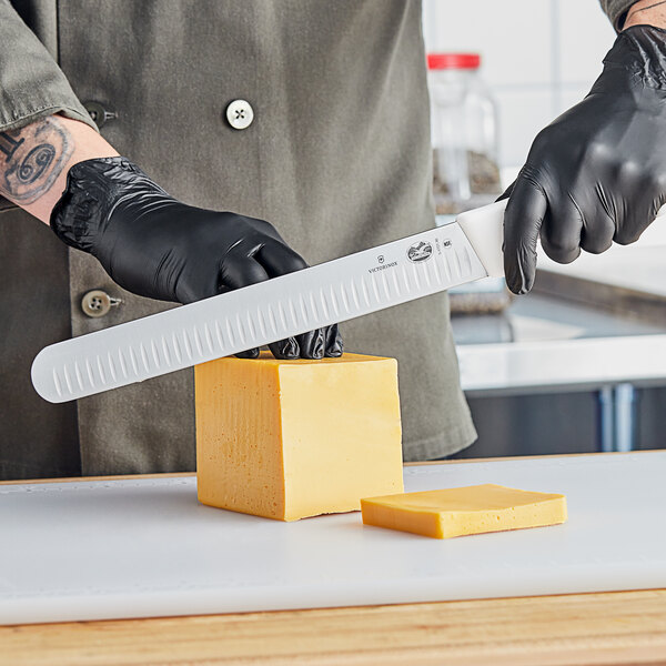 A person's hand in black gloves using a Victorinox slicing knife to cut cheese.