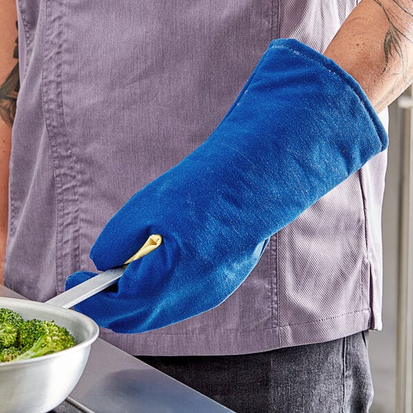 A person wearing a Tucker Safety blue oven mitt holding a pan of broccoli.