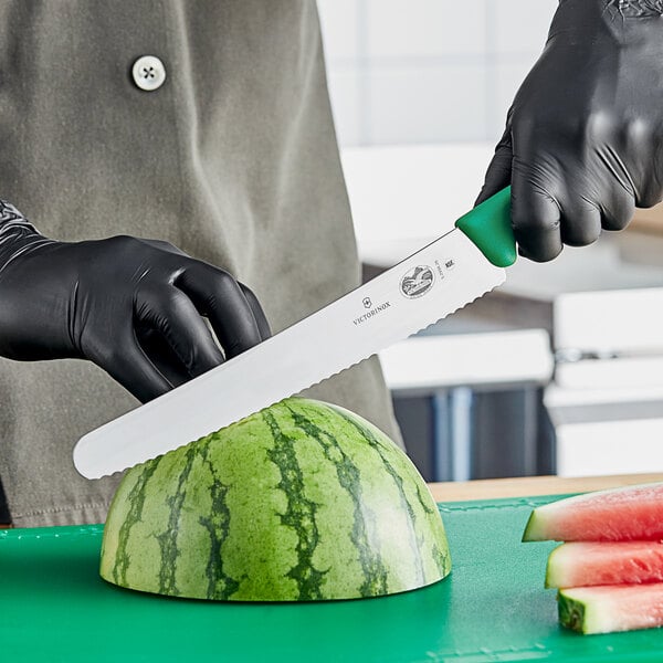 A person wearing a black glove uses a Victorinox curved serrated bread knife to cut a watermelon.