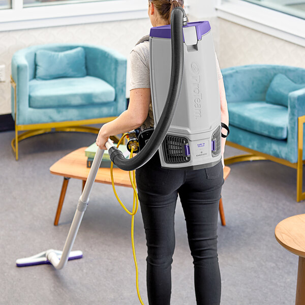 A woman using a white ProTeam backpack vacuum cleaner with a black hose to vacuum a room.