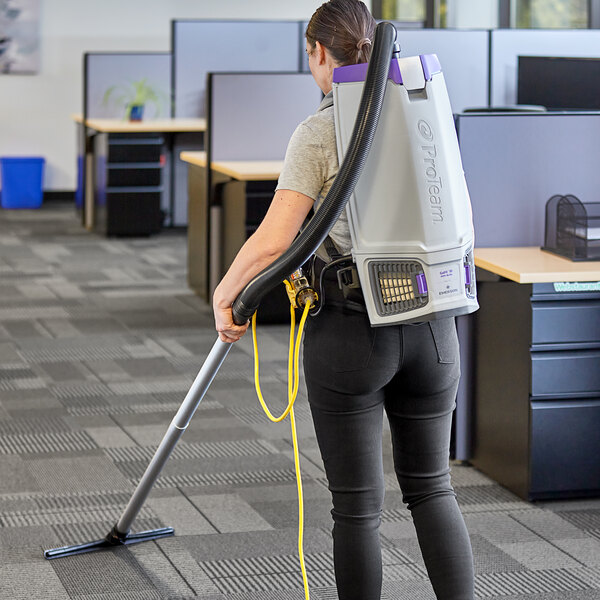 A woman using a ProTeam GoFit backpack vacuum to vacuum carpet in a room.