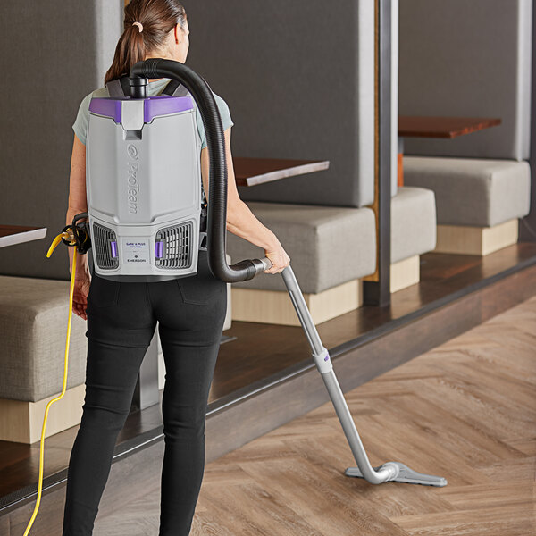 A woman using a ProTeam backpack vacuum to clean a room.