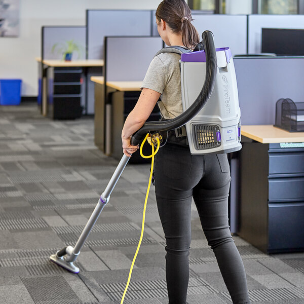 A woman using a ProTeam GoFit backpack vacuum to clean a carpet.