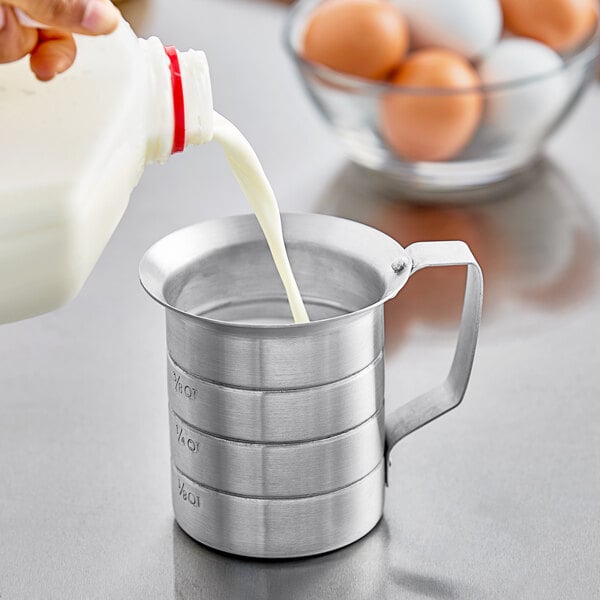 A person pouring white liquid into a Choice aluminum measuring cup.