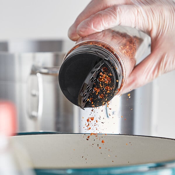 A person in gloves pouring seasoning into a container using a black dual-flapper spice lid with 7 holes.