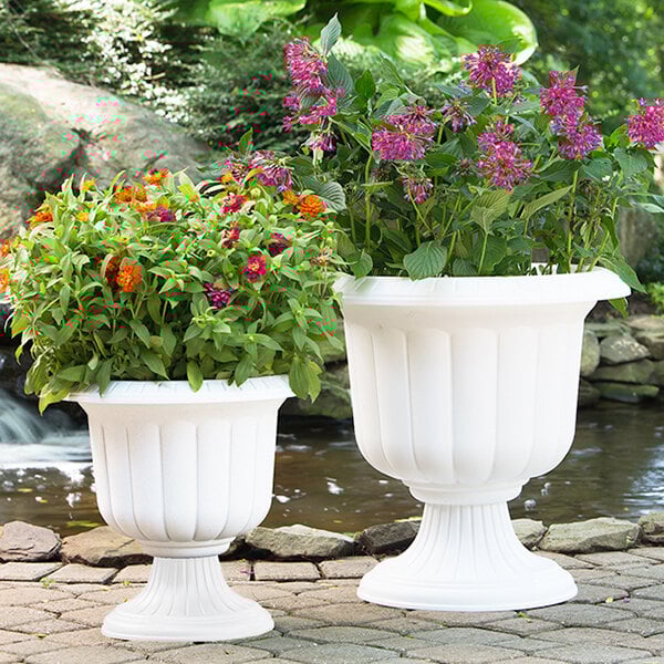 Two white Novelty Manufacturing Co. urn planters with flowers in them.