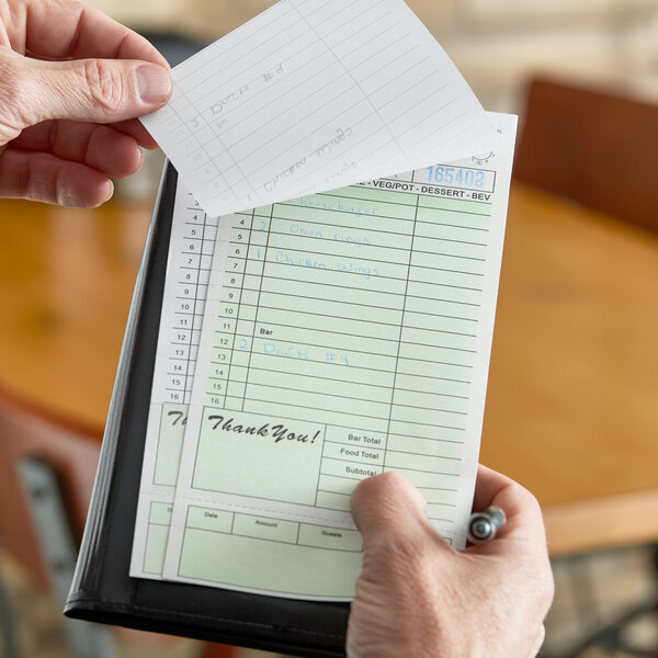 A green and white carbonless guest check with a bottom guest receipt being held in a restaurant setting.