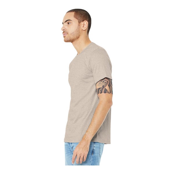 A man wearing a Bella + Canvas heather dust t-shirt and jeans with a tattoo on his arm.