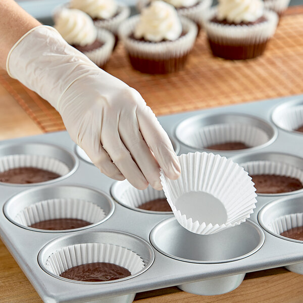 A person wearing gloves is putting a cupcake with white frosting into a white fluted baking cup in a muffin tin.