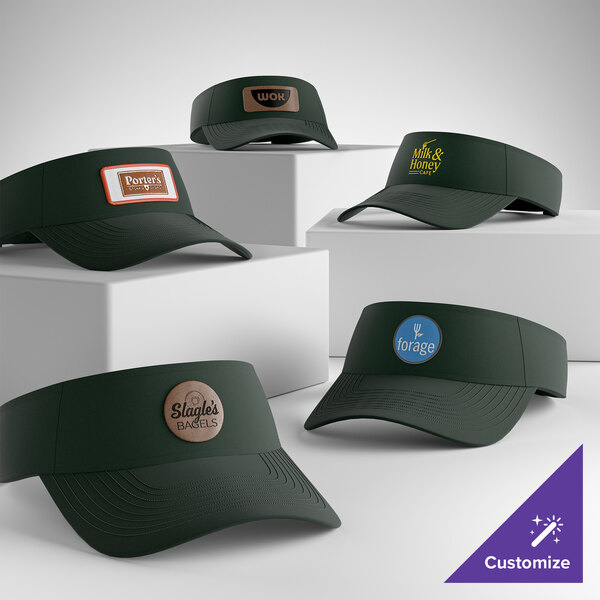 A collection of dark green Port & Company CP45 customizable hunter visors displayed on white blocks, each featuring different custom logos.