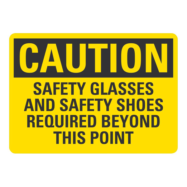 A yellow sign with black text that reads "Caution Safety Glasses and Safety Shoes Required Beyond This Point"