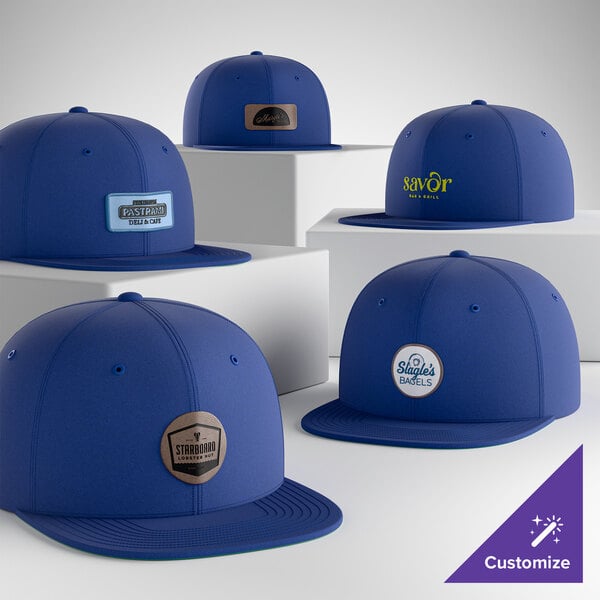 A collection of customizable royal blue 6-panel flat bill snapback caps with various logo designs on the front.
