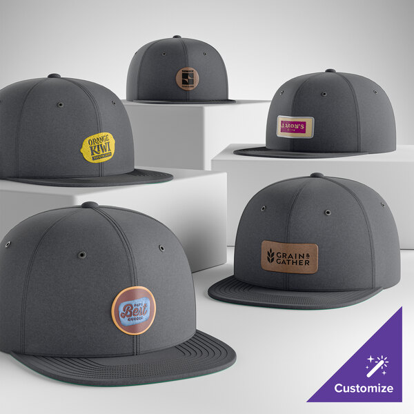 A collection of customizable graphite 6-panel flat bill snapback caps with various logo patches on the front.