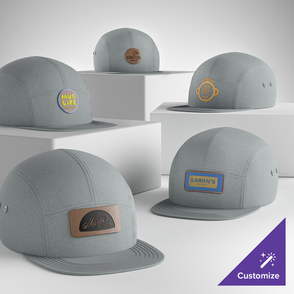 A collection of customizable deep smoke gray 7-panel flat bill camper caps with various logo patches on the front.