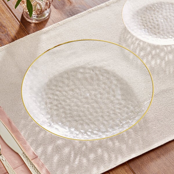 A table setting with a Visions clear plastic coupe plate with a gold rim, knife and fork on a placemat.