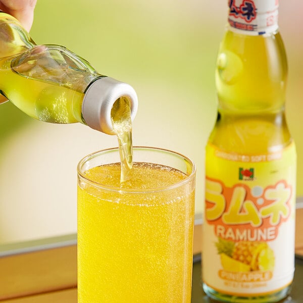 A hand pouring Hana Ramune Pineapple Soda from a yellow bottle into a glass.