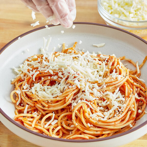 A person's gloved hand sprinkling Plant Ahead vegan shredded mozzarella cheese on a bowl of spaghetti.