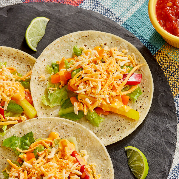 A group of three tacos with Plant Ahead vegan shredded cheese and vegetables on a black plate.