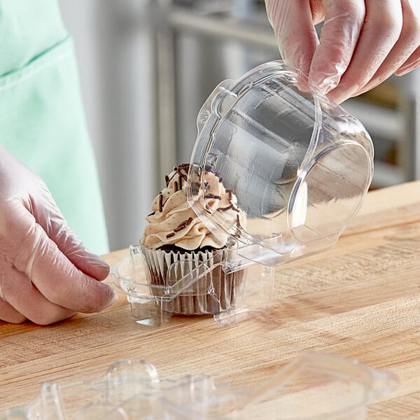 A gloved hand placing a cupcake with frosting and sprinkles in a clear plastic container.