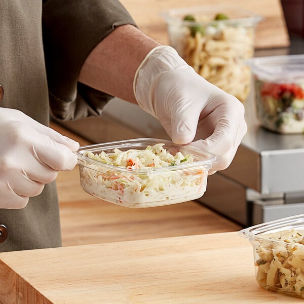 A person in gloves holding a Good Natured clear PLA square deli container of food.