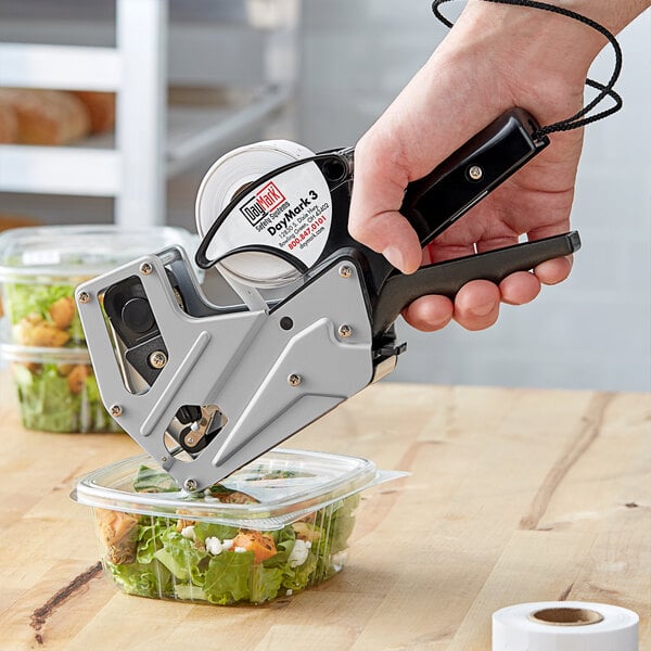 A hand using a DayMark Speedy Mark 3 metal date coder to label a plastic container of salad.