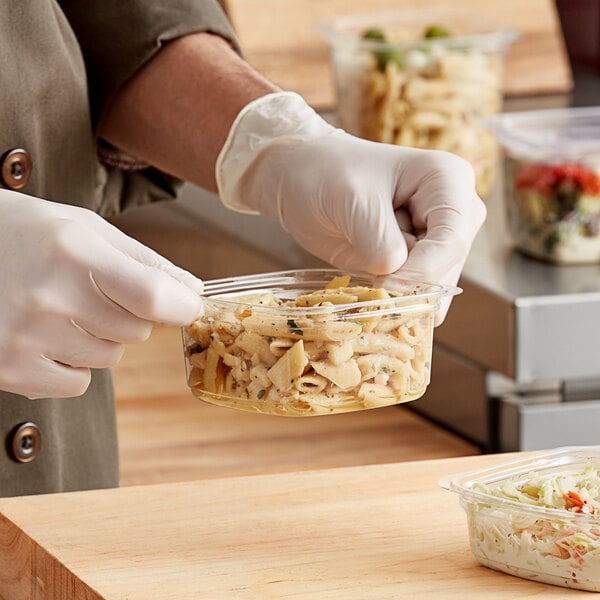 A gloved hand holding a Good Natured clear plastic deli container of pasta on a wood surface.