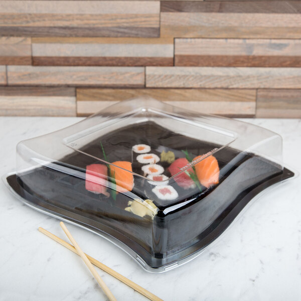 A clear plastic dome lid on a clear plastic container with sushi inside and chopsticks on the table.