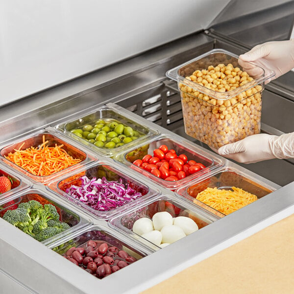 A person putting shredded carrots into a clear plastic food pan on a salad bar counter.