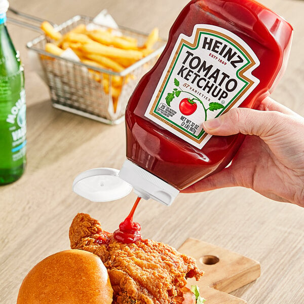 A hand using a Heinz Ketchup Upside Down Squeeze Bottle to pour ketchup onto a chicken sandwich.