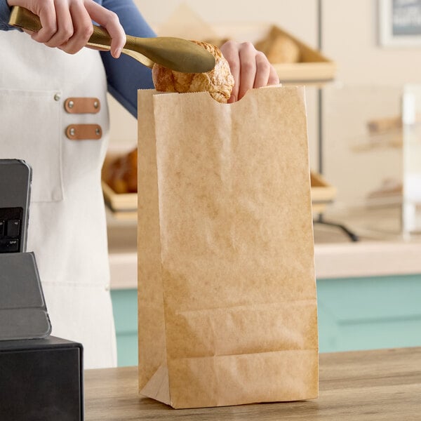 A large natural kraft waxed paper bag being filled with a pastry using tongs.