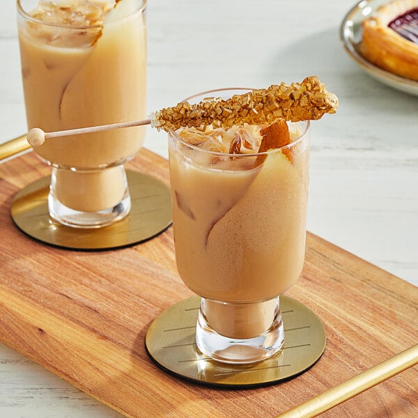 Two glasses of brown liquid with a Fanale Golden Pear Rock Sugar syrup stick on a wooden table.