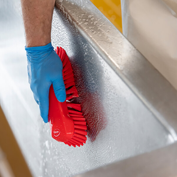 A person wearing blue gloves using a Vikan red scrub brush to clean a metal surface.