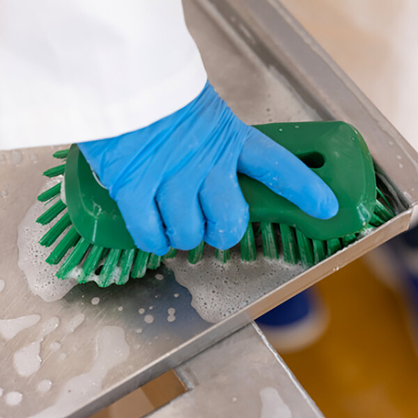 A person in blue gloves cleaning a metal surface with a Vikan green scrub brush.