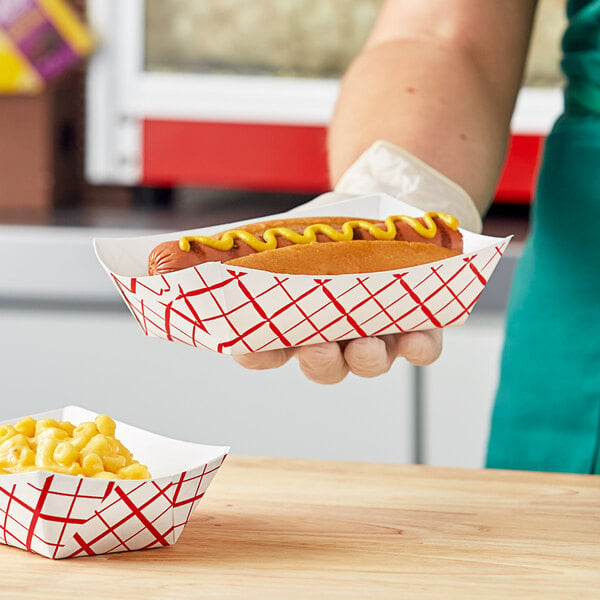 A person holding a Southern Champion red checkered paper food tray with a hot dog and a bowl of macaroni and cheese.